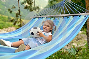 A little girl lies in a hammock with a ball and smiles.