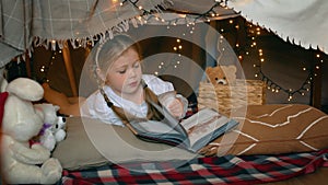 A little girl lies and reads a book in a tent of blankets and pillows in the bedroom. Children