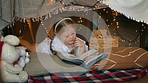 A little girl lies and reads a book in a tent of blankets and pillows in the bedroom. Children