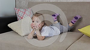 A little girl lies on the sofa among the pillows and moves her legs. Home recreation for children