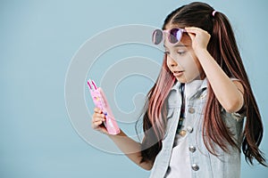 Little girl lifting her sunglasses, looking at her phone. Over blue background
