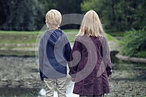 Little Girl with light hair And Boy In Blue Jacket Standing Back On Bank Of River And looking on the park