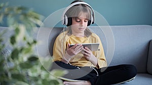 Little girl listening to music with headphones and digital tablet while sitting on sofa at home.