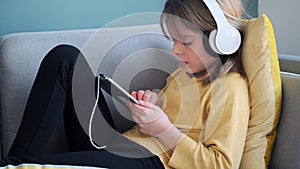 Little girl listening to music with headphones and digital tablet while sitting on sofa at home.