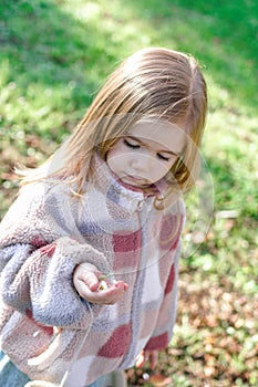 A little girl with long blonde hair in a pink and white checkered jacket kneels in a grassy field.