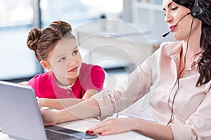 Little girl looking at busy mother in headset working with laptop