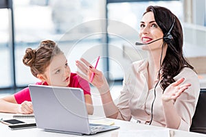 Little girl looking at happy mother in headset working in office