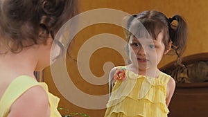 A little girl is looking in the mirror. A beautiful girl with tails on her head leans in front of the mirror. A child in a yellow