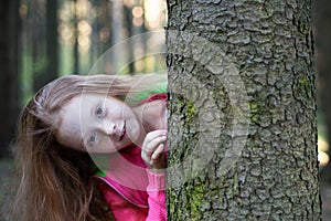 Little girl looks from behind a tree in the forest