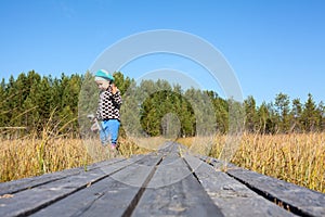Little girl looks for in grass standing near wooden planks of pathway passing through the swampland, copy space photo
