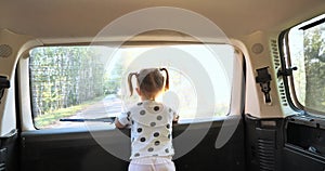 A little girl looks out the back window of a car while driving through the forest. The child stands near the rear window