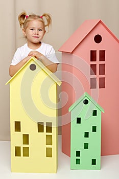 A little girl looks out from behind a toy wooden house.