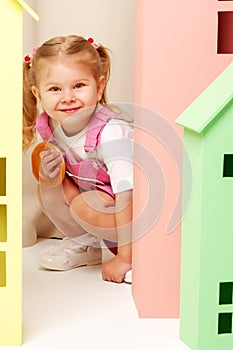 A little girl looks out from behind a toy wooden house.