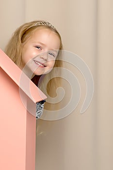 A little girl looks out from behind a toy wooden house.