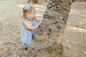 A little girl looks out from behind the trunk of a palm tree on the beach sand, childlike spontaneity,