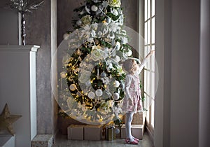 A little girl looks out of a large window near a Christmas tree. Waiting for a miracle, Christmas white decor in the living room o