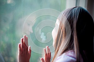 Little girl looks out the window at her reflection in the glass on a summer day