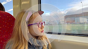 A little girl looks out the window of the train. A 7-year-old sad child girl rides a train and looks out the window. 4K