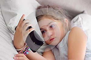 Little girl lying on the bed with her smartphone, being bored, watching alone