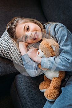 Little girl lying on couch with teddy bear and smiling at camera