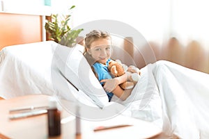 Little girl lying in hospital bed with teddy bear and smiling at camera