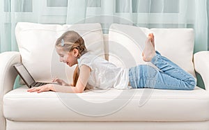 Little girl lying on sofa looks at notebook and laughs