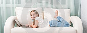 Little girl lying on sofa looks at notebook and laughs