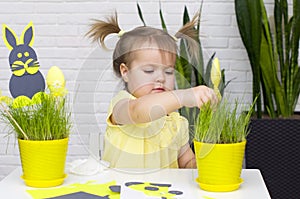 A little girl makes  Easter decorations, arranges handmade decorative elements in a pot of green grass, a Easter bunny and eggs