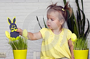 A little girl makes  Easter decorations, arranges handmade decorative elements in a pot of green grass, a Easter bunny and eggs