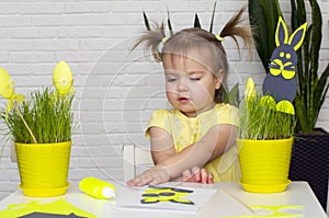 A little girl makes  Easter decorations, arranges handmade decorative elements in a pot of green grass, a Easter bunny and eggs