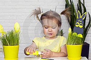 A little girl makes  Easter decorations, arranges handmade decorative elements in a pot of green grass, a Easter bunny and eggs