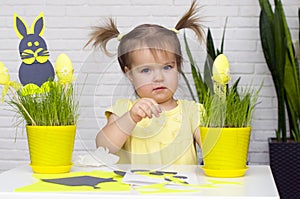 A little girl makes  Easter decorations, arranges handmade decorative elements in a pot of green grass, a Easter bunny and eggs