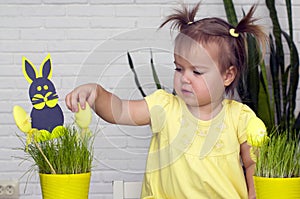 A little girl makes  Easter decorations, arranges handmade decorative elements in a pot of green grass, a Easter bunny and eggs