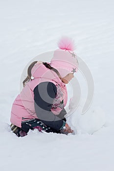 Little girl making big snow ball for constructing snow man in park in daytime with ground covered with snow in