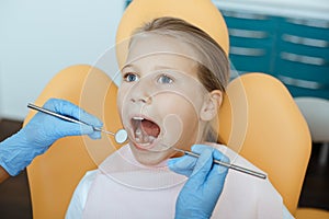 Little girl in medical chair opens her mouth, dentist in rubber gloves with tools treats teeth or put filling