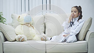 Little girl in medical coat is talking in the stethoscope. White teddy-bear in medical mask is sitting near her.
