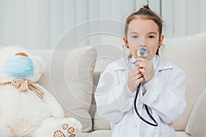 Little girl in medical coat is talking in the stethoscope. White teddy-bear in medical mask is sitting near her.