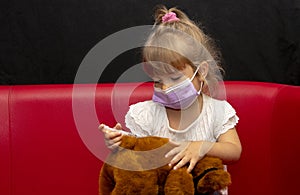 A little girl in a medical mask holds a syringe in her hands and gives injections to a teddy bear sitting on the couch. Concept: p