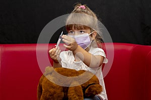 A little girl in a medical mask holds a syringe in her hands and gives injections to a teddy bear sitting on the couch. Concept: p