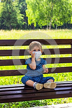 A little girl in a medical mask sits alone on a bench in a summer Park.