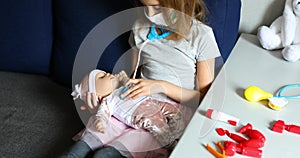 A little girl in medical mask sits on the sofa, plays a doctor, treats to the doll
