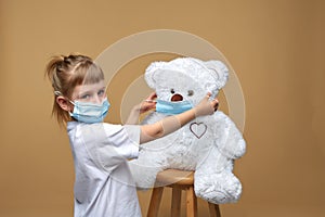 A little girl in a medical mask stands and puts a medical mask on her toy teddy bear