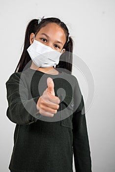 Little girl with medical mask with the thumb up with gesture of hope and victory on a white background. Concept of positivism photo