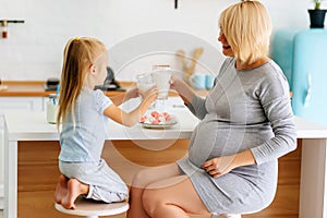 A little girl with mom, a young girl with a pregnant blonde, is poured and drink milk in the kitchen at the table