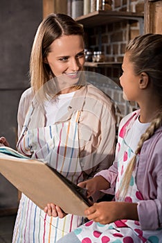 little girl and mother in aprons looking at each other while reading cookery book together photo