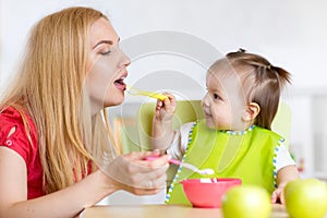 Little girl and mother with baby food feeding each other, sitting at table in nursery