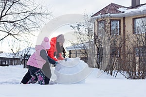 Little girl with mother rolling with high efforts snow balls for making snowman on cottage backyard in evening with