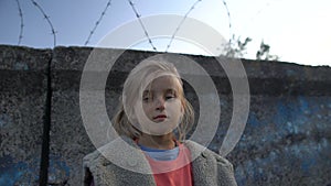 Little girl with muddy face standing at barbed wire, war child in refugee camp