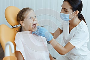 Little girl open her mouth for treatment, young female doctor in white protective mask and gloves