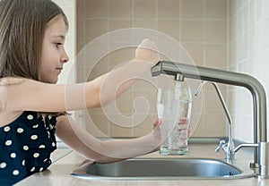 Little girl open a water tap with her hand holding a transparent glass. Kitchen faucet. Filling cup beverage. Pouring fresh drink